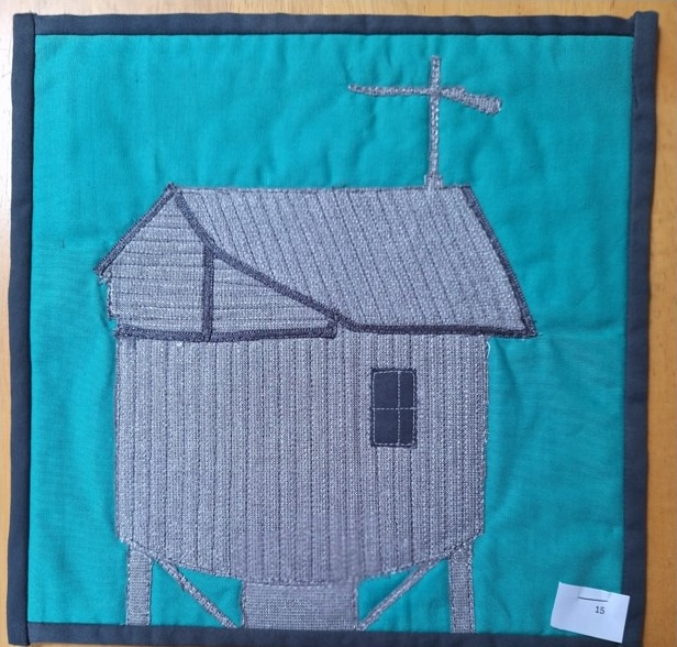 A square quilt with a turquoise background and a black border. The quilt features a gray house with a slanted roof and a small window. The house is on stilts and has an antenna on top