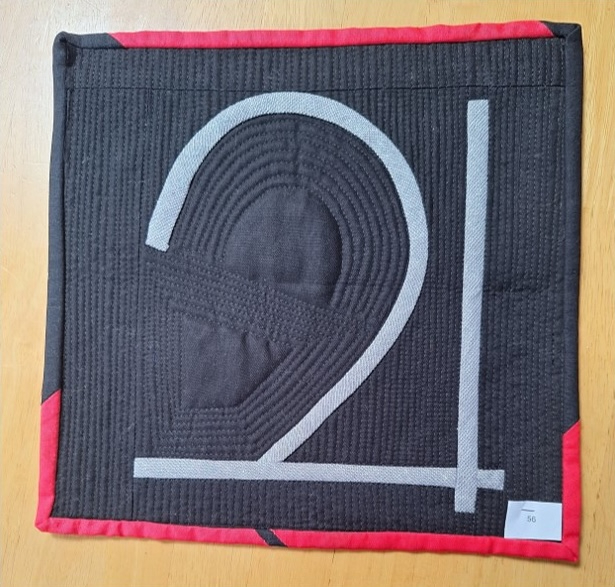 A square quilt with a black background and a red border. The quilt features the number "21" in white, with the "2" having a layered, circular design. The background is quilted with a linear pattern that echoes the focal point.