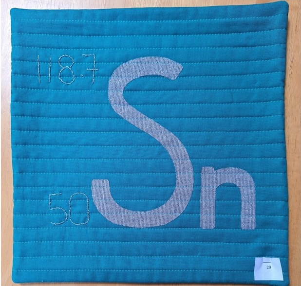 A square quilt with a blue background and a gray border. The quilt features the chemical symbol for Tin, "Sn," with its atomic number (50) and atomic weight (118.7) displayed above and below. The background is quilted with vertical lines.