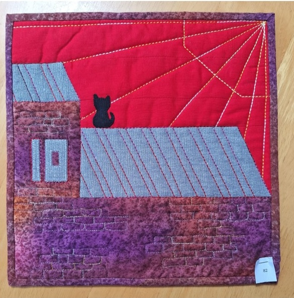 A square quilt with a red background and a black border. The quilt features a black cat silhouette sitting on a gray roof. The background is quilted with radiating lines, suggesting a sunset. The house has a purple, textured base.