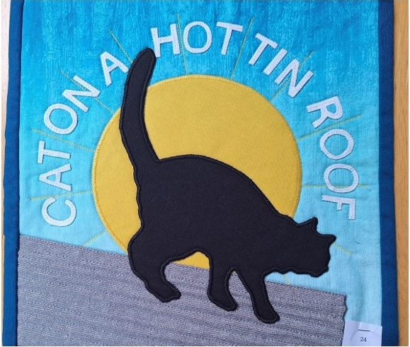 A square quilt with a blue background. The quilt features a black cat silhouette walking on a grey rooftop. A yellow sun with the words "CAT ON A HOT TIN ROOF" surrounds the cat. The roof is quilted with a linear pattern to mimic a corrugated tin roof.