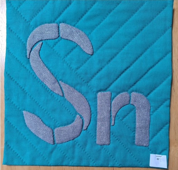 A square quilt with a turquoise background and a gray border. The quilt features the letters "Sn" in gray, with the "S" made up of smaller, gray shapes. The background is quilted with a diagonal line pattern.