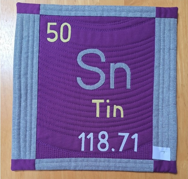 A square quilt with a purple background and a gray border. The quilt features the chemical element Tin, with its atomic number (50), symbol (Sn), and atomic weight (118.71) displayed on a purple background. The background is quilted with a wavy pattern.