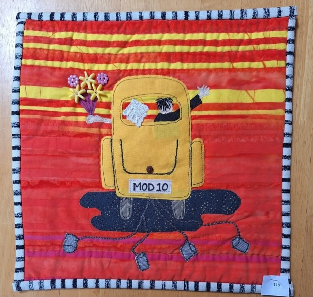 A square quilt with a red background and a black and white striped border. The quilt features a yellow car with a bride and groom inside, driving through a puddle. The car has the number "MOD 10" on the side.