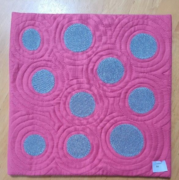 A square quilt with a pink background and a pink border. The quilt features a pattern of gray circles surrounded by concentric pink circles. The background is quilted with a circular pattern.