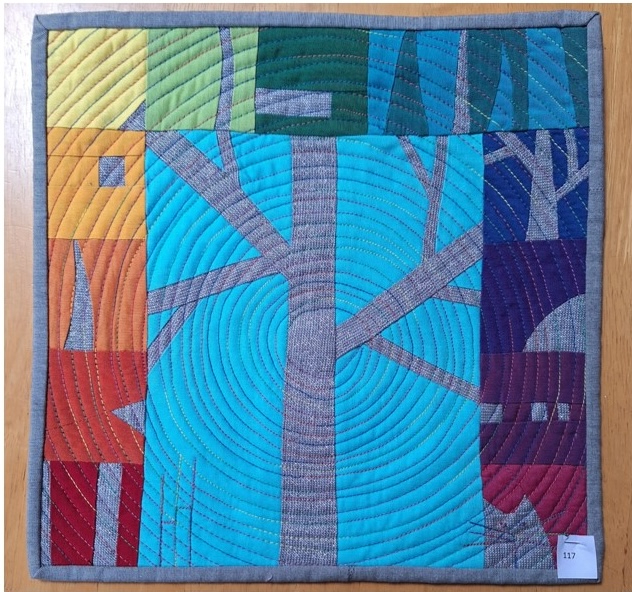 A square quilt with a gray background and a gray border. The quilt features a rainbow-coloured background with a gray tree in the centre. The tree has branches and leaves, and there are houses on either side. The background is quilted with a circular pattern.