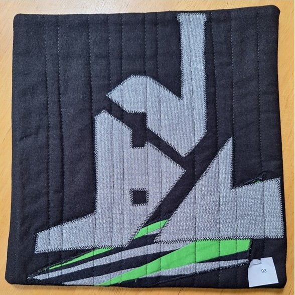 A square quilt with a black background and a black border. The quilt features a gray, abstract shape that resembles a factory or a building. The background is quilted with a linear pattern, and there are green stripes at the bottom.