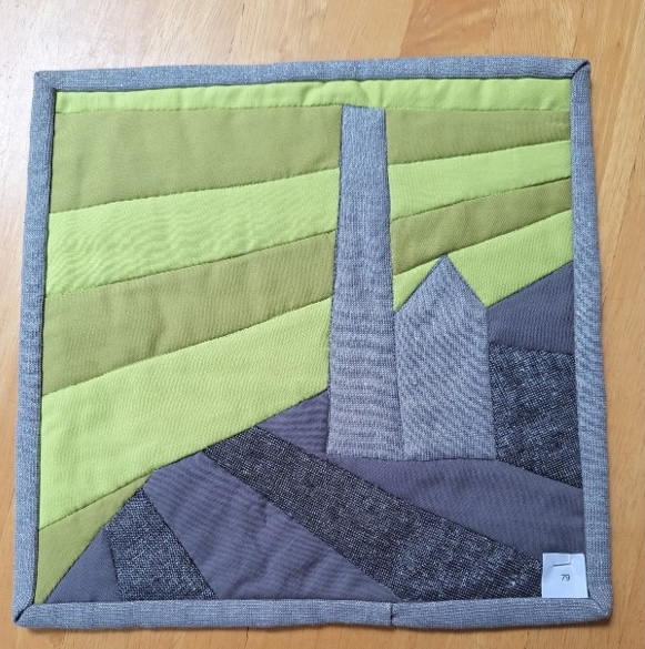 A square quilt with a gray background and a gray border. The quilt features a landscape scene with a gray building on a hill. The sky is depicted with shades of green fabric.