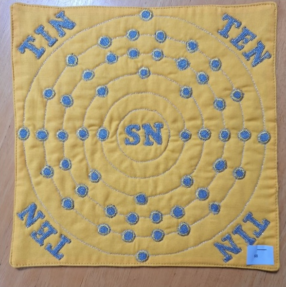 A square quilt with a yellow background and a gray border. The quilt features a stylized representation of the Tin atom, with the symbol "Sn" in the centre and the word "TIN" repeated around the edges. The background is quilted with a circular pattern.