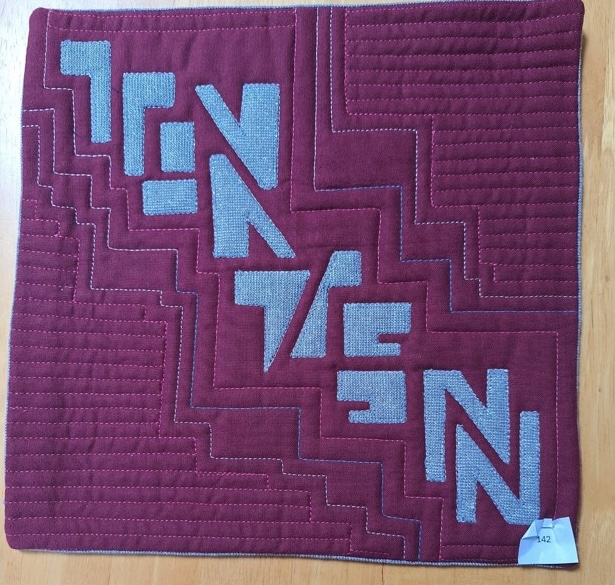 A square quilt with a red background and a red border. The word "TEN" is repeated four times in gray letters, arranged in a diagonal pattern. The background is quilted with a linear and steps pattern.