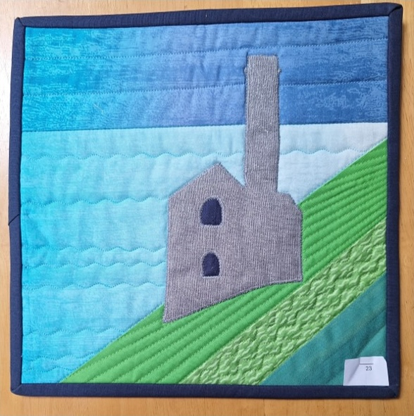 A square quilt with a blue background and a blue border. The quilt features a gray building with a chimney, perched on a green hill overlooking a blue body of water. The sky is depicted with shades of blue fabric