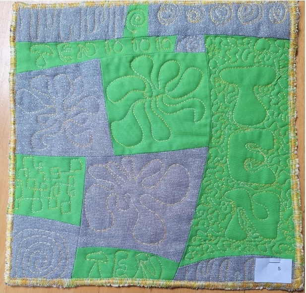 A square quilt with a gray background and a yellow border. The quilt features a patchwork design with squares and rectangles in green and gray. Some of the shapes have embroidered patterns or textures.