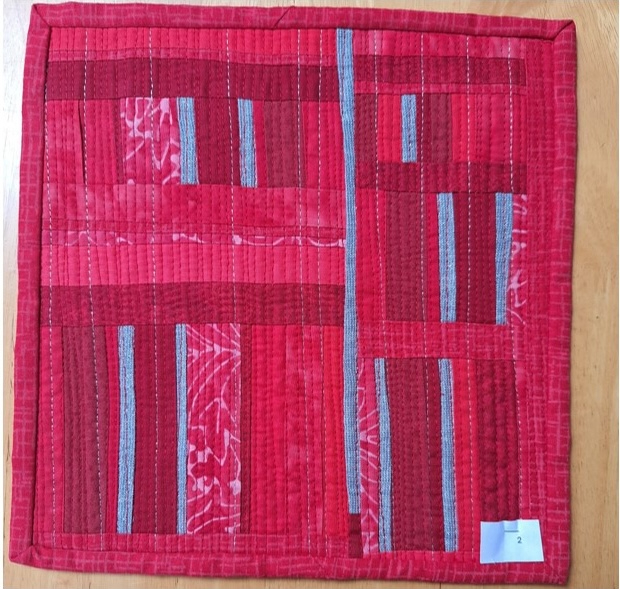 A square quilt with a red background and a red border. The quilt features a pattern of vertical and horizontal lines in various shades of red, with some areas of white. The background is quilted with a linear pattern.
