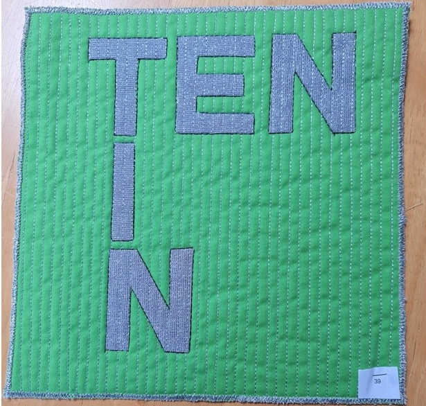 A square quilt with a green background and a gray border. The quilt features the word "TEN" and the letter "N" stacked vertically, both made of gray fabric. The background is quilted with vertical lines.