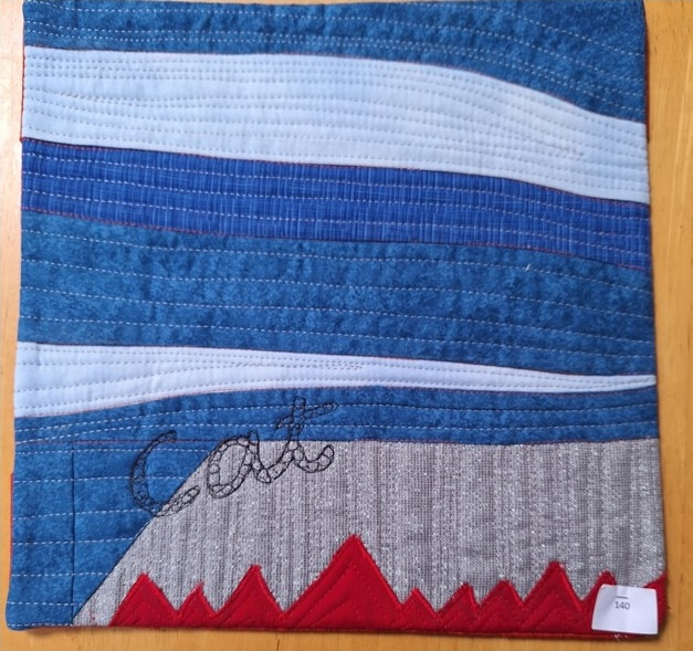 A square quilt with a blue background and a red border. The quilt features a landscape with horizontal stripes representing the sky and a mountain range at the bottom. The word "CAT" is embroidered on the mountain range. The background is quilted with a linear pattern.