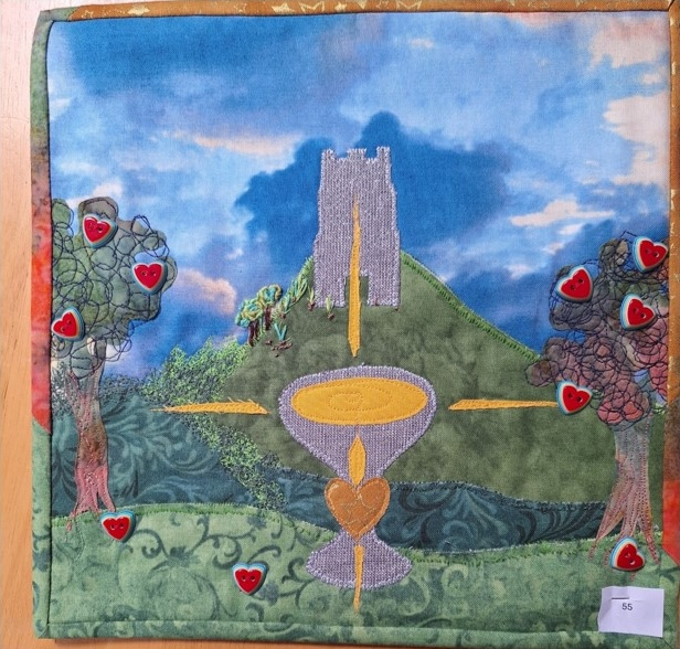 A square quilt with a colourful landscape scene. The quilt features a hill with a castle on top, two trees on either side, and a golden chalice in the centre. The sky is blue with white clouds.