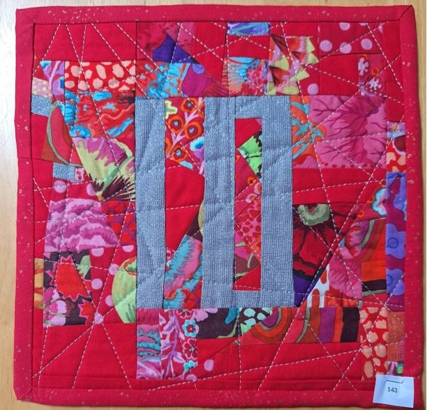 A square quilt with a red background. The quilt features the number "11" in a gray, stylized font. The background is filled with a patchwork of colourful fabric pieces with various patterns.