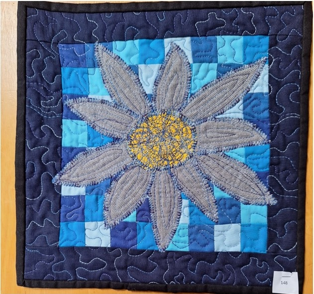 A square quilt with a blue background and a black border. The quilt features a large, gray flower with a yellow centre. The flower is surrounded by a checkered pattern of blue and white squares. The background is quilted with a stipple pattern.