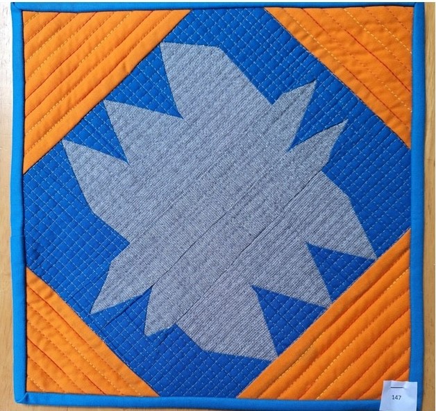 A square quilt with a blue background and an orange border. The quilt features a gray, star-shaped design in the centre. The background is quilted with a linear pattern.