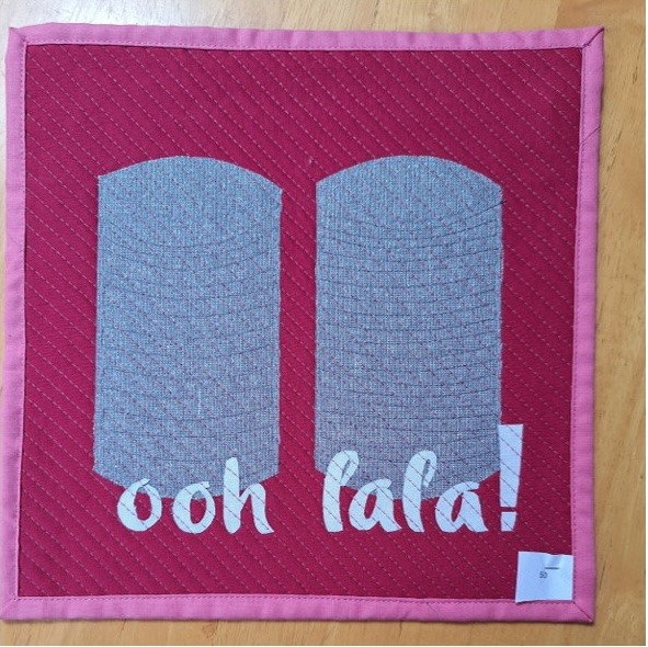 A square quilt with a red background and a pink border. The quilt features two gray, cylindrical shapes representing tin cans, with the phrase "ooh la la!" appliquéd below.