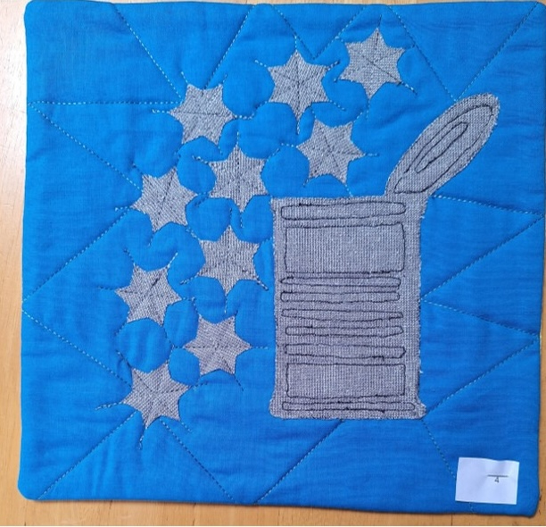 A square quilt with a blue background and a gray border. The quilt features a gray tin can with stars spilling out of it. The background is quilted with a triangular pattern.
