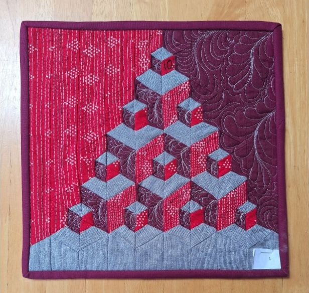 A square quilt with a red background and a red border. The quilt features a pyramid-shaped design made up of overlapping, gray cubes. The background is quilted with a linear pattern and a swirling pattern.