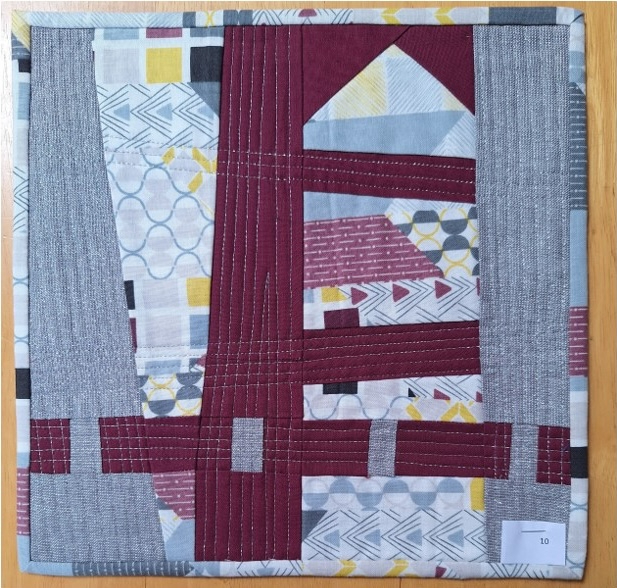 A square quilt with a gray background and a red border. The quilt features a patchwork design with rectangles and triangles in various colours and patterns, including gray, red, yellow, and white. The background is quilted with a linear pattern.