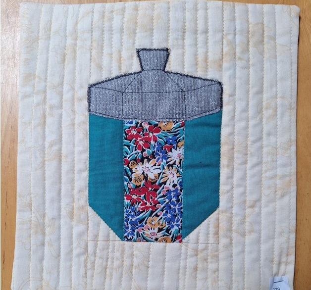 A square quilt with a cream background. The quilt features a colourful dreidel, a traditional spinning top used in the Jewish holiday of Hanukkah. The dreidel is made of various fabrics with different patterns. The background is quilted with a linear pattern.