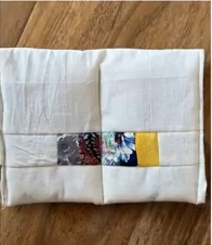 A white quilted needle case with a colorful patchwork strip in the center. The strip features various patterns and colors, including floral and geometric designs.