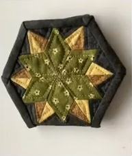 A hexagonal quilted needle case with a star-shaped design in shades of green and brown. The star is made up of smaller triangles and has a quilted texture. The case has a dark brown border.