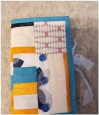 A blue quilted needle container with a patchwork design. The patchwork features various patterns and colors, including blue, yellow, white, and gray. The container has buttons and a blue ribbon tie.