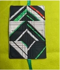 A black quilted needle case with a geometric patchwork design. The patchwork features black, white, green, and red triangles arranged in a symmetrical pattern. The case has a green ribbon tie.