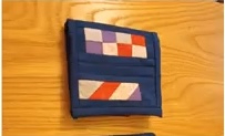A blue quilted needle case with a patchwork design. The patchwork features squares and stripes in red, white, and purple.