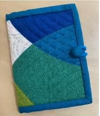 A blue quilted needle case with a patchwork design. The patchwork features shades of blue, green, and white fabric, arranged in a curved pattern. The case has a blue button closure.