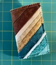 A brown and blue quilted needle container with a patchwork design. The container is made up of strips of fabric in various shades of brown, beige, and blue, arranged in a diagonal pattern.