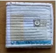A white quilted needle case with a patchwork design. The patchwork features various shades of white and a small blue section. The case has a button closure.