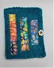 A teal quilted needle case with a colorful patchwork design. The patchwork features strips of fabric in various shades of blue, purple, and yellow. The case has a small button closure.