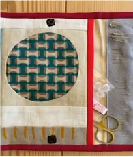 A red and white quilted needle case with a circular window showing a teal fabric with a geometric pattern. The case has pockets for pins, scissors, and other sewing tools.