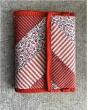 A red quilted needle container with a patchwork design. The patchwork features a combination of red and white stripes and a paisley pattern. The container has a flap that closes with a snap.