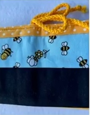 A blue and yellow quilted needle case with a bee theme. The case has a black bottom section and a blue top section with a pattern of bees and flowers. It closes with a yellow drawstring.