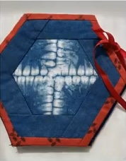A hexagonal quilted needle case with a dark blue background and a red border. The central design features a white, X-shaped pattern with a blue and white floral motif in the center. The case has a red ribbon tie.