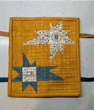 A yellow quilted needle case with two star-shaped designs. One star is made of blue fabric with a sewing machine design in the center, and the other is made of white fabric with a different design. The background is quilted with a linear pattern.