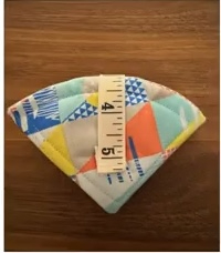 A colorful, quilted needle case in the shape of a fan. The case is made of various fabrics with geometric patterns and bright colors like blue, yellow, orange, and white. A measuring tape fastens the case together.