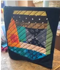 A black quilted needle case with a colorful patchwork design. The patchwork features various shapes and colors, including red, orange, yellow, green, and blue. The case has a quilted texture and a black border.