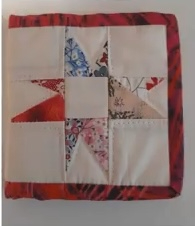 A red quilted needle container with a patchwork design. The patchwork features a star shape made up of red, white, and blue fabric pieces. The container has a red border.