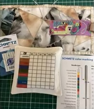 A photo of a sewing kit on a green cutting mat. The kit includes a quilted needle case with a colorful design, a small pouch with a chart inside, and a packet of sewing needles.