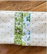 A white quilted needle case with a vertical strip of green fabric featuring floral patterns. The background fabric has a subtle polka dot pattern.