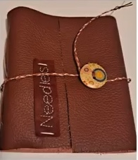 A brown leather needle case with a decorative button and the word "Needles" embroidered on the front. The case is closed with a string tie.