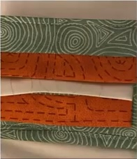 A green and orange quilted needle case with a geometric pattern. The case has a white interior with a pocket for needles.
