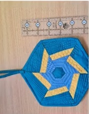 A blue and yellow quilted needle case shaped like a hexagon. The case features a star-like design in the center with a ruler placed next to it for scale.